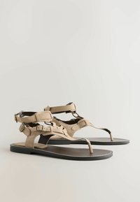 Sandales plates beiges en daim style gladiateur avec semelles noires, brides autour de la cheville et du pied, et boucles réglables sur un fond uni.