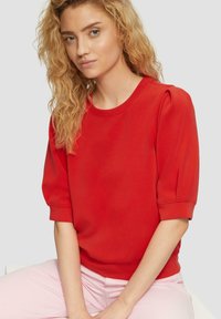 Top rosso a maniche corte con scollatura tonda e maniche a sbuffo, realizzato in tessuto liscio. Il modello indossa pantaloni rosa chiaro, seduto su uno sfondo grigio.