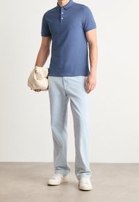 Polo bleu à trois boutons, associé à un jean bleu clair et des baskets blanches. Modèle tenant un sac beige. Tenue simple et décontractée.