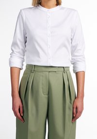 Persona che indossa una camicia bianca a maniche lunghe con le maniche arrotolate e pantaloni a vita alta plissettati di colore verde oliva.