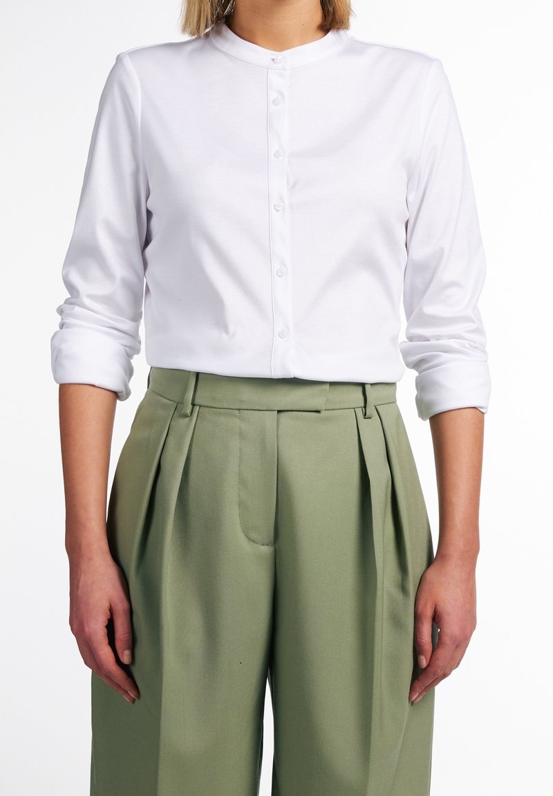 Persona che indossa una camicia bianca a maniche lunghe con le maniche arrotolate e pantaloni a vita alta plissettati di colore verde oliva.