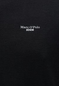 Crni materijal s bijelim tekstom "Marc O’Polo DENIM" centriranim na površini.