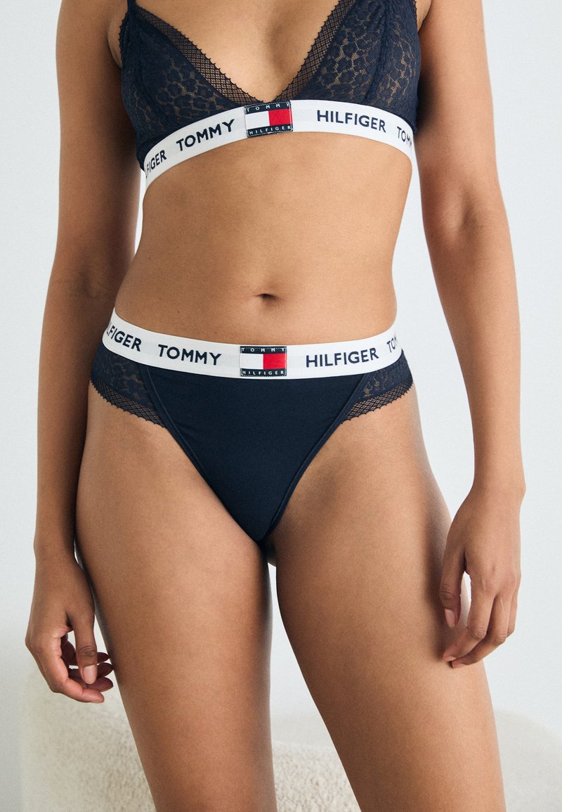 Zwarte bralette met kant en een witte elastische band met het logo "TOMMY HILFIGER". Bijpassende string met kantaccenten en een vergelijkbare tailleband.