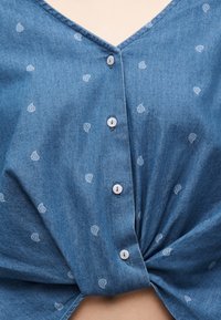Μπλε υφασμάτινο πουκάμισο με μικρά λευκά παϊέρι μοτίβα, δεμένο σε κόμπο στο κάτω κέντρο.