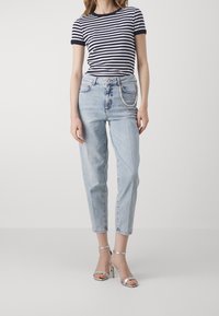 Kvinna iklädd en svart och vit randig t-shirt, ljusblå högmidjade jeans med en kedja, och silverfärgade högklackade sandaler står.