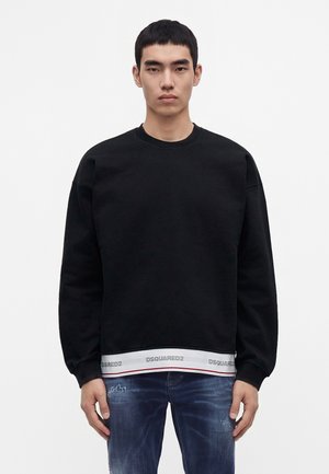 Jeune homme aux cheveux courts et noirs porte un sweat-shirt noir sur une chemise blanche avec le logo DSQUARED2, assorti à un jean bleu délavé.