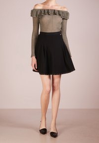 Olivenfarbener, rippierter Off-Shoulder-Top mit Rüschensaum, kombiniert mit einem schwarzen A-Linien-Rock, der goldene Nieten am Saum hat. Schwarze spitze Ballerinas.