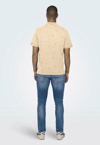 Beige kurzärmliges Hemd mit kleinen, dunklen Mustern. Kombiniert mit blauen Jeans. Model zeigt nach hinten und trägt Sneaker.