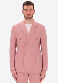 Uomo che indossa un blazer doppiopetto rosa pallido e pantaloni abbinati, in piedi davanti a uno sfondo bianco, con lo sguardo rivolto in avanti.