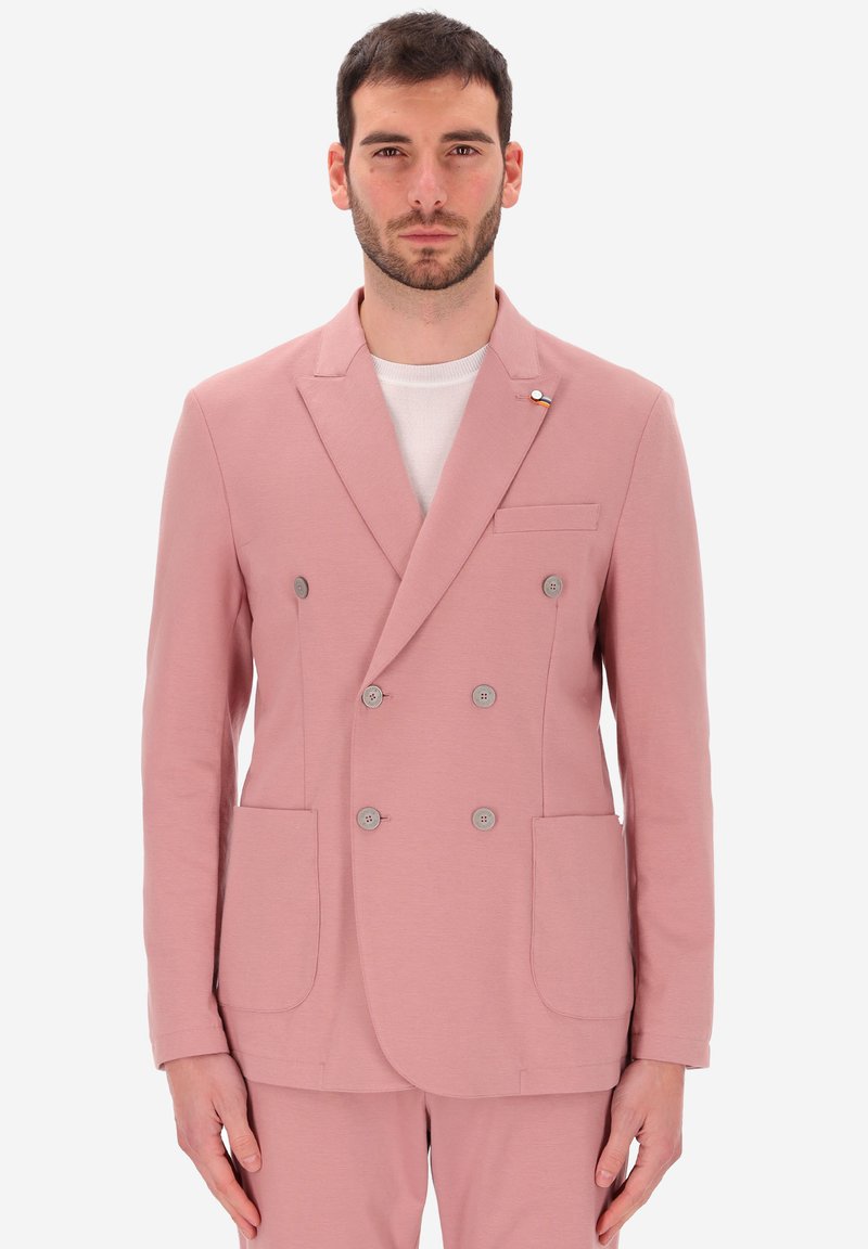 Uomo che indossa un blazer doppiopetto rosa pallido e pantaloni abbinati, in piedi davanti a uno sfondo bianco, con lo sguardo rivolto in avanti.