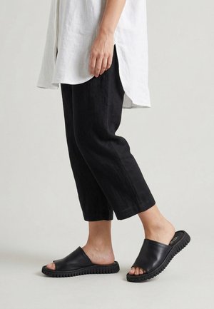 Personne portant un pantalon noir raccourci, des sandales noires sans brides et une longue chemise blanche, debout sur un fond clair uni.