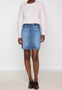 Kvinna iklädd en vit stickad tröja, högmidjad blå denimkjol med knappar fram, och mörkbruna loafers, stående mot en vit bakgrund.