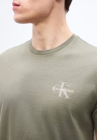 Νεαρός άνδρας που φοράει ανοιχτό ελιάς t-shirt Calvin Klein με στρογγυλή λαιμόκοψη, εμφανίζοντας το άνω μέρος του στήθους και μερικό προφίλ του προσώπου.