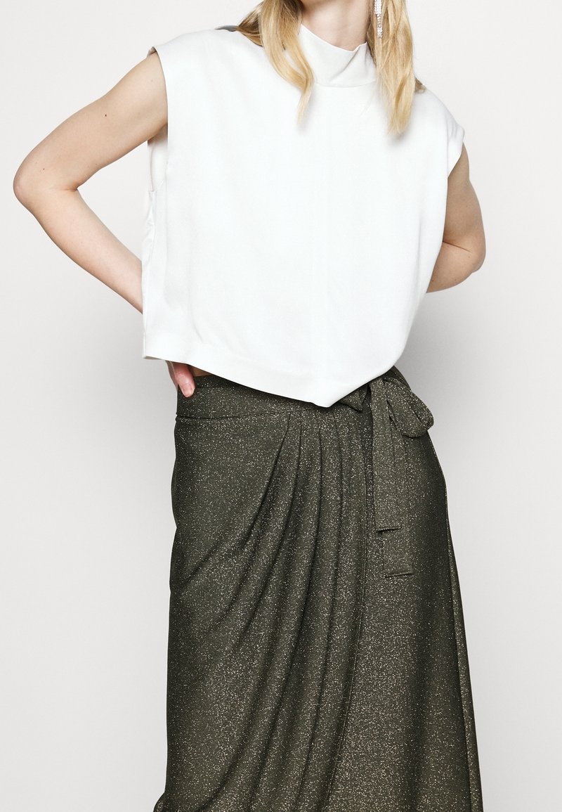 Femme portant un haut blanc sans manches et une jupe pailletée vert foncé taille haute avec une ceinture nouée, posant les mains sur les hanches.