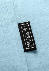 Hellblaues Gewebe mit einer glatten Textur, das ein schwarzes rechteckiges Etikett mit weißem Text "SPITZBUB" und einem kleinen Logo in der Ecke zeigt.