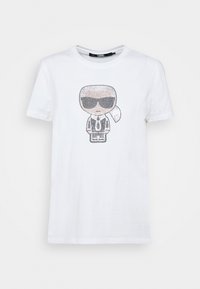 Valkoinen puuvillapaita, jossa on graafinen kuva sarjakuvahahmosta, joka käyttää aurinkolaseja ja pukua, koristeltuna mustilla ja hopeisilla strasseilla.