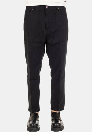 Uomo che indossa pantaloni neri slim-fit e scarpe slip-on in pelle nera su uno sfondo bianco semplice.