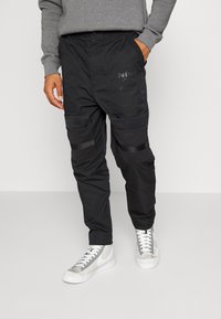 Pantalones joggers negros con cintura elástica, con detalles de malla y múltiples bolsillos. Combinados con una camiseta de manga larga gris y zapatillas blancas.