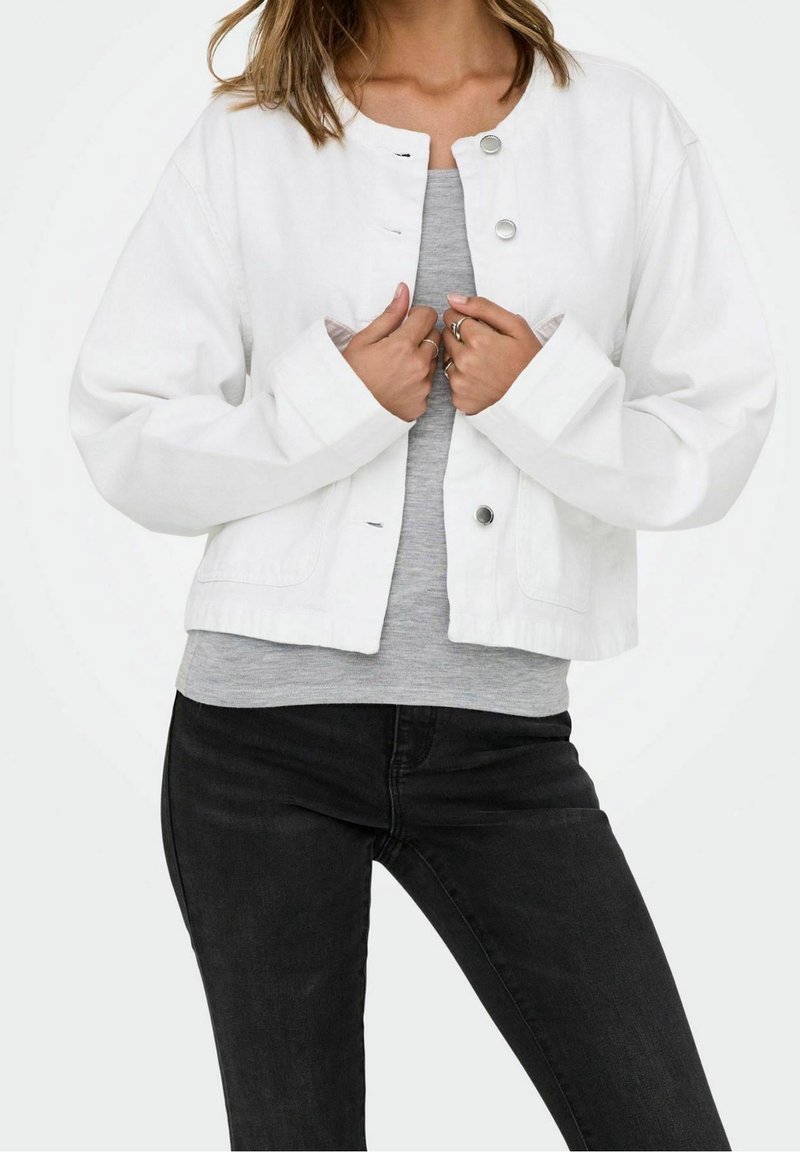 Femme portant une veste courte blanche à boutons sur une chemise grise et un pantalon noir, tenant la veste près de la poitrine.