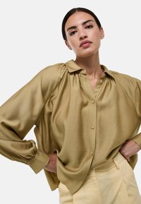 Blouse en satin vert olive avec un devant à boutons, coupe décontractée, et un léger plissage aux épaules. Manches longues avec poignets à boutons.