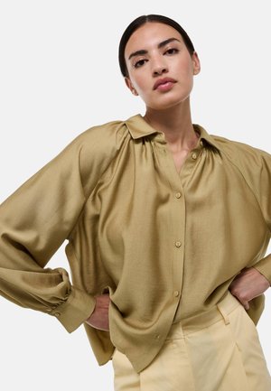 Blouse en satin vert olive avec un devant à boutons, coupe décontractée, et un léger plissage aux épaules. Manches longues avec poignets à boutons.