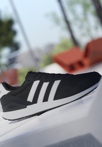 Sneaker Adidas nero con strisce bianche e suola bianca appoggiato su una superficie bianca, sfondo esterno sfocato con vegetazione e mobili rossi.