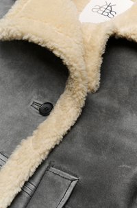 Grijze suede jas met een crèmekleurige shearling kraag, voorzien van een zwarte knop en opgestikt zakje, met een zachte textuur en strakke lijnen.