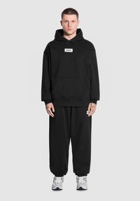 Czarna bluza z kapturem, z przednią kieszenią i naszywką logo, w połączeniu z czarnymi dresami. Materiał wydaje się miękki; strój ma luźny krój i ściągacze przy mankietach.