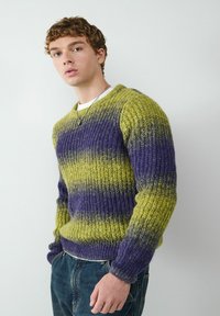 Gebreide trui met een gestructureerd patroon in groene en paarse strepen, met een ronde halslijn en geribde manchetten, gecombineerd met donkerblauwe jeans.