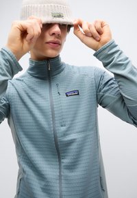 Άτομο που φοράει ανοιχτό γκρι ζακέτα Patagonia με φερμουάρ και μπεζ σκουφάκι, κατεβάζοντας το σκουφάκι πάνω από τα μάτια του, μπροστά σε απλό φόντο.