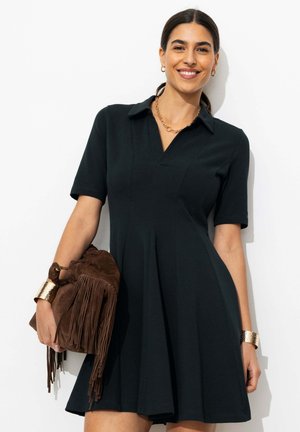 Femme en robe noire à manches courtes tenant un sac en daim marron à franges, portant des boucles d'oreilles créoles dorées, un collier chaîne épais et de larges bracelets dorés.
