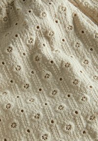 Cremfarbene Stoff mit kunstvoller Stickerei und floralen Mustern sowie kleinen Ausschnitten, die ein strukturiertes Gitterdesign bilden. Weicher Look.
