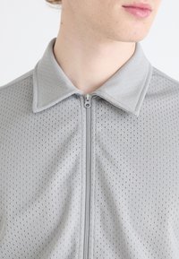 Grå zip-up skjorta med perforerad textur, som har en krage och subtila sömnadsdetaljer. Närbild av den övre halvan.
