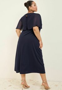 Navyblaues Kleid mit einem fließenden Cape-Rückenteil, einem Schlüssel-Ausschnitt und einer taillierten Passform. Kombiniert mit metallischen Riemchen-Heels.