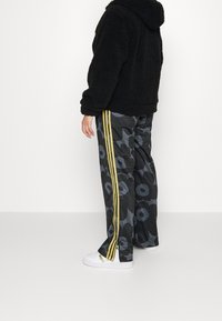 Sudadera negra de felpa, pantalones deportivos grises con patrón y franjas amarillas a los lados, y zapatillas blancas. Los pantalones presentan un diseño floral y un corte holgado.