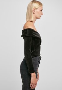 Sorte kordfløyels off-shoulder topp med lange ermer og en tettsittende design, kombinert med mørke jeans. Jevn tekstur og strukturert silhuett.