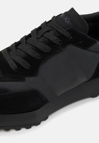 Black sneaker featuring a mix of suede and synthetic materials, with prominent laces, textured panels, and a branded side accent.