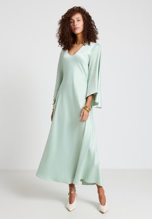 Woman with curly hair wearing a mint green satin long-sleeve dress, gold bracelets, and white pointed heels, standing on a plain white background.