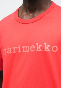 Czerwona bawełniana koszulka z okrągłym dekoltem i jasnym napisem "marimekko" na piersi. Gładka tekstura i dopasowany krój.