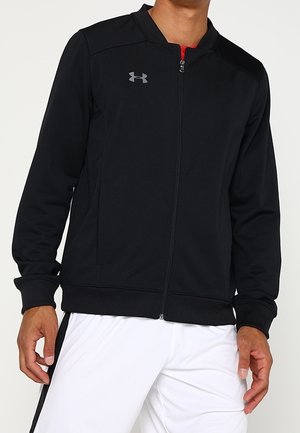 Mann trägt eine schwarze Sportjacke mit Reißverschluss und kleinem grauem Logo auf der Brust und weiße Hose mit schwarzem Streifen.