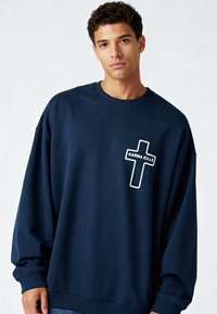 Jeune homme portant un sweat-shirt ample bleu foncé avec une croix blanche et le texte « KARMA KILLS » sur la poitrine, devant un fond uni clair.
