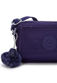 Fioletowa torebka crossbody marki Kipling z regulowanym paskiem oraz przymocowanym do metalowego pierścienia pluszowym breloczkiem w kształcie goryla.