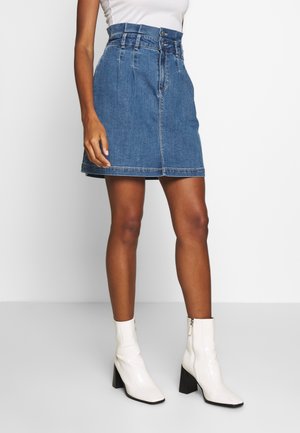 Kvinde iført en blåt højtaljet denimnederdel med knapper foran og hvide ankelstøvler med hæl mod en ensfarvet baggrund.