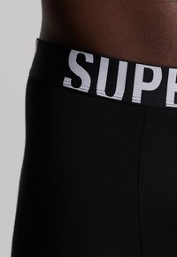 Cintura negra con "SUPER" en letras blancas en negrita en la parte superior de pantalones ajustados negros. Textura suave con un ligero brillo.
