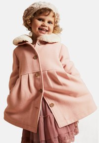 Lyserød uldfrakke med en faux fur-krave, der har store knapper og et flagrende design. Kombineret med en lagdelt lyserød tylnederdel.
