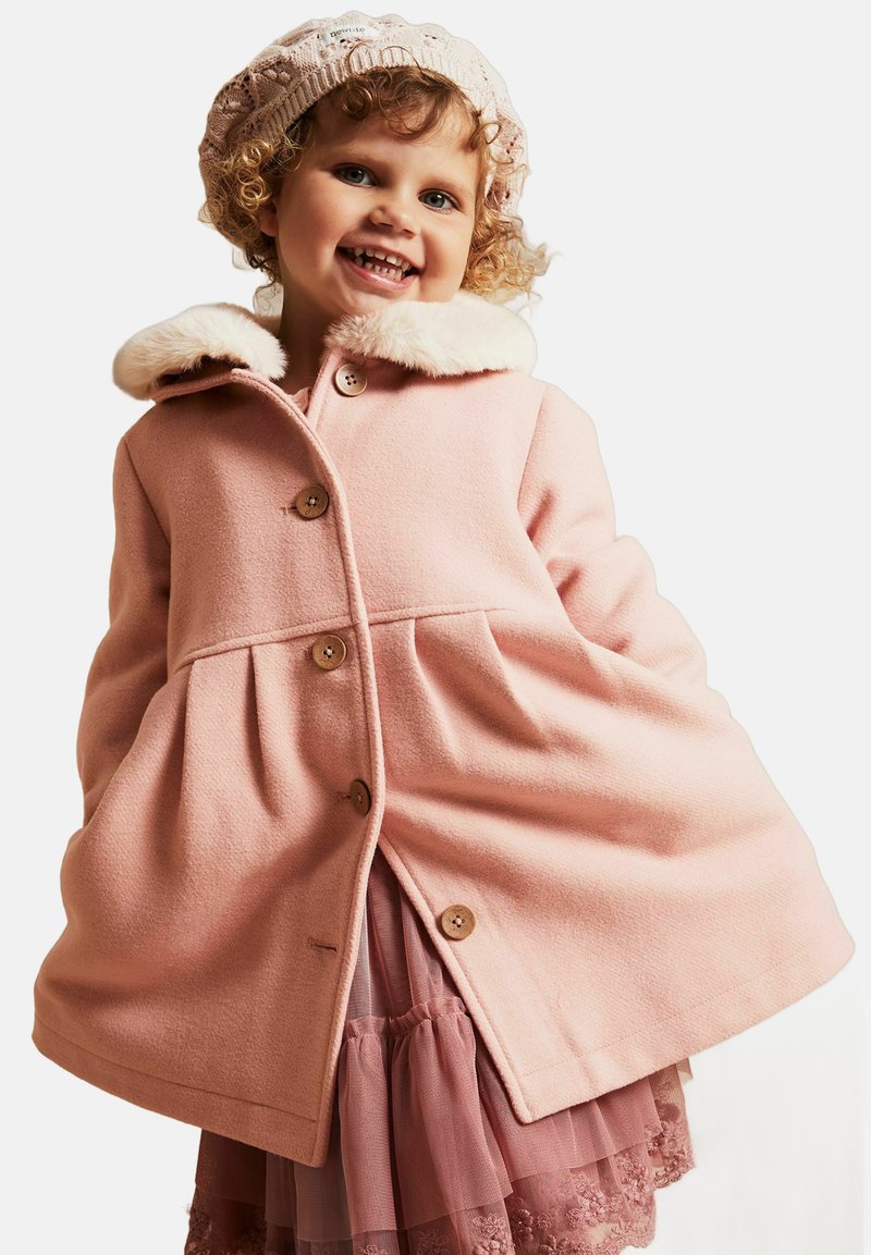 Lyserød uldfrakke med en faux fur-krave, der har store knapper og et flagrende design. Kombineret med en lagdelt lyserød tylnederdel.