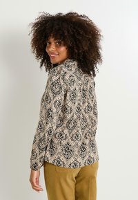 Blusa de manga larga con un patrón de paisley en negro y beige, con una textura suave y un cuello con solapa. Combinada con pantalones de color verde oliva.