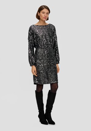 Robe noire à paillettes avec un décolleté bateau, des manches longues et une taille élastique. Portée avec des bottes noires à hauteur de genou, mettant en avant une coupe slim et une texture brillante.