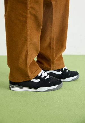 Person som har på seg brune kordfløyelbukser og svarte Rowley-sneakers med hvite lisser, står på grønt gulv.