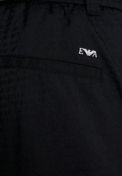 Mustmustest kangad, millel on peene logo muster ja hõbedane embleem "EVA" tagataskus. Sile tekstuur ja kohandatud lõige.